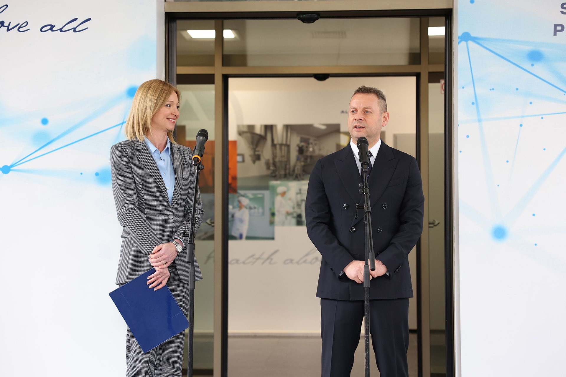 A man and woman in business attire standing and talking in a modern office lobby with glass doors.