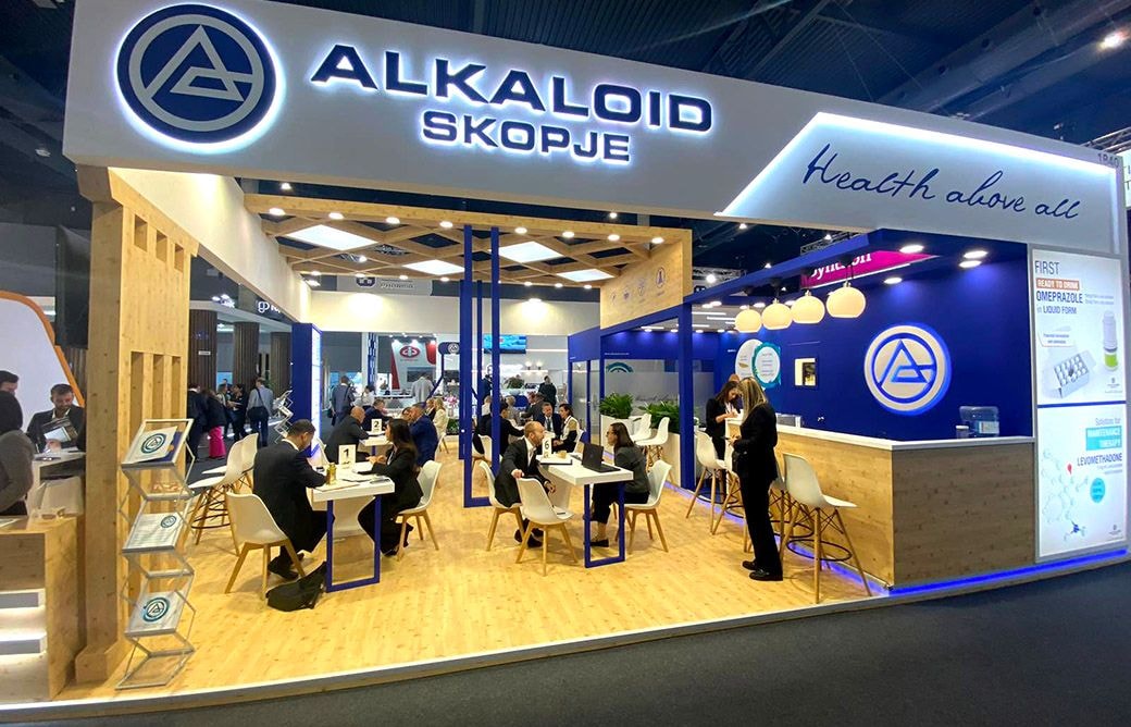 Exhibition booth with "ALKALOID SKOPJE" and "Health. Idea. Life." signage, showing people seated at tables and standing in conversation at a trade fair.