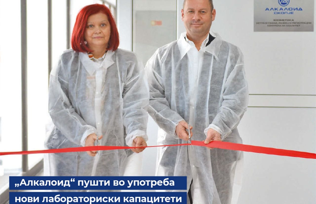 Two people in white cleanroom coveralls, gloves, and hairnets cutting a ribbon to mark the opening of a laboratory or production facility.