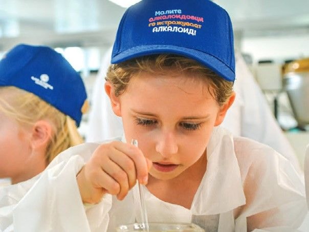 Close-up of a child in a blue baseball cap with a white logo, wearing a white lab-style coat, using a pipette or dropper during a hands-on science activity.